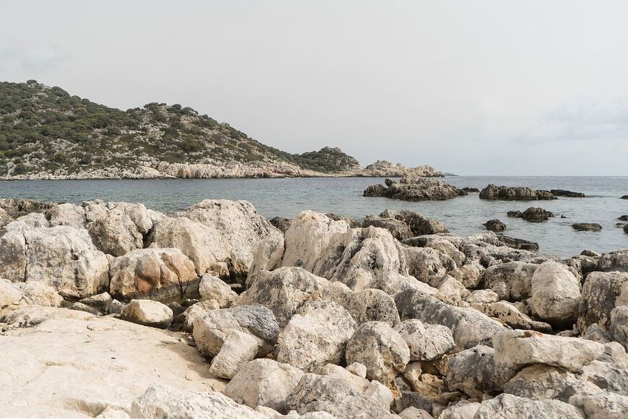 Pale, weathered limestone boulders on the shore of Kekova Island with a calm bay and a scrub-covered hillside behind them.