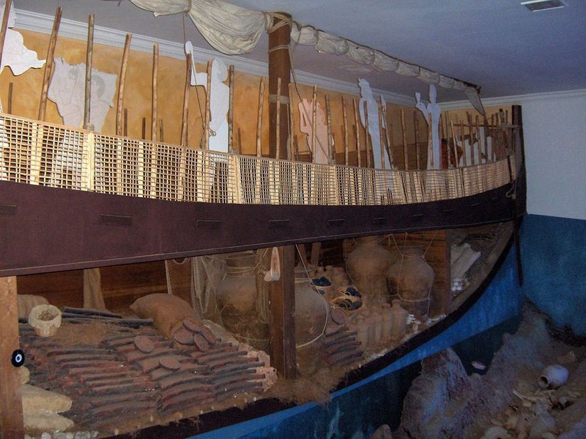 A cutaway reconstruction of an ancient wooden ship in a museum hall, with a woven side rail, mast and sail, and jars and metal ingots stacked inside the hull.