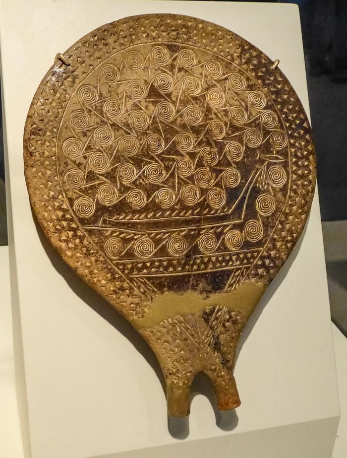 A round clay vessel with a short handle, its base incised with a ship and fish surrounded by spiral and linear patterns, shown on a museum stand.