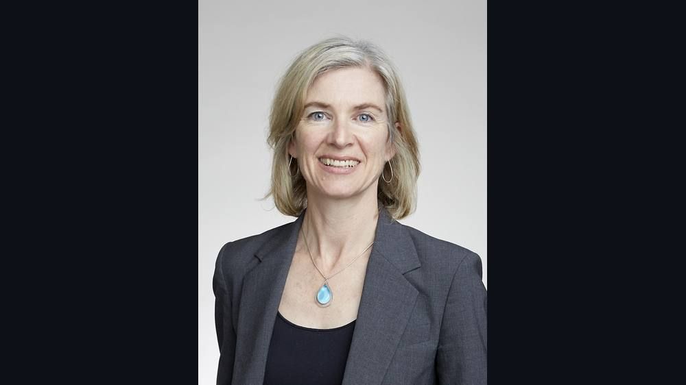 A portrait of a woman with blonde hair in a gray blazer with a pendant necklace.