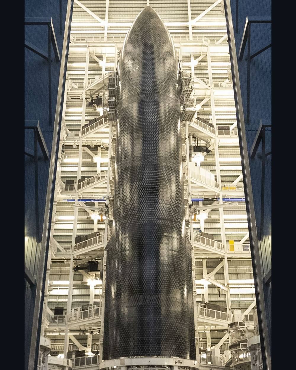 A dark tile-covered Starship hull stands inside a tall steel work structure with several levels of platforms.
