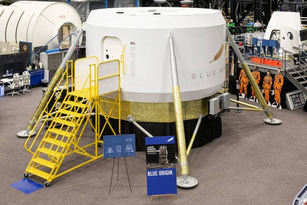 A white cylindrical crew-cabin mock-up with a stairway stands in a large NASA training hall.
