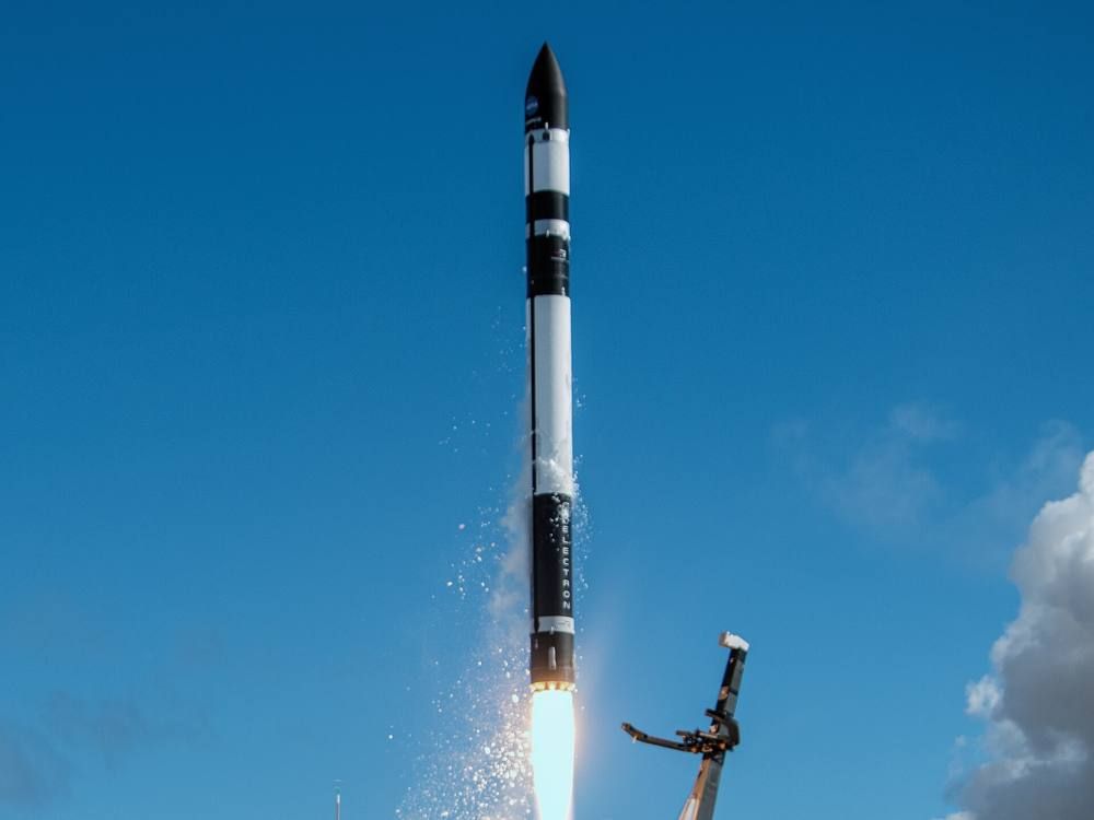 A slim black and white rocket climbs against a bright blue sky with a plume of exhaust at its base.