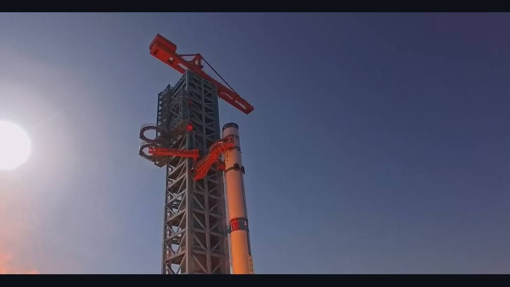 A tall white and red rocket stands beside a steel launch tower with bright orange flame and smoke at the left under a hazy blue sky.
