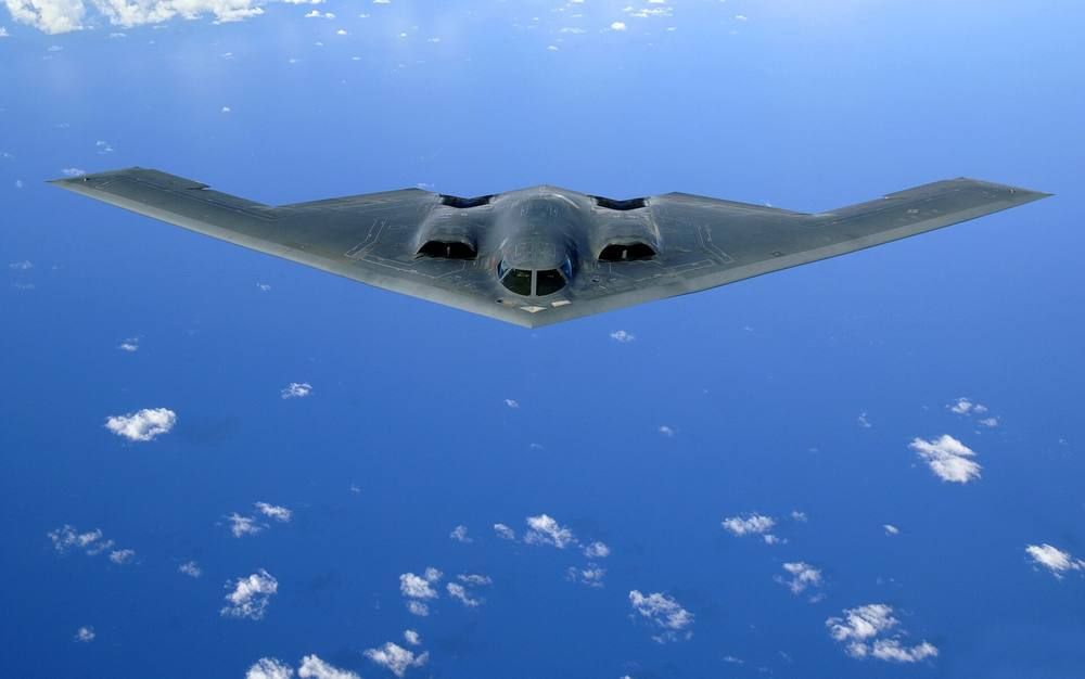 A dark gray triangular flying-wing bomber seen head-on over blue ocean with scattered white clouds below.